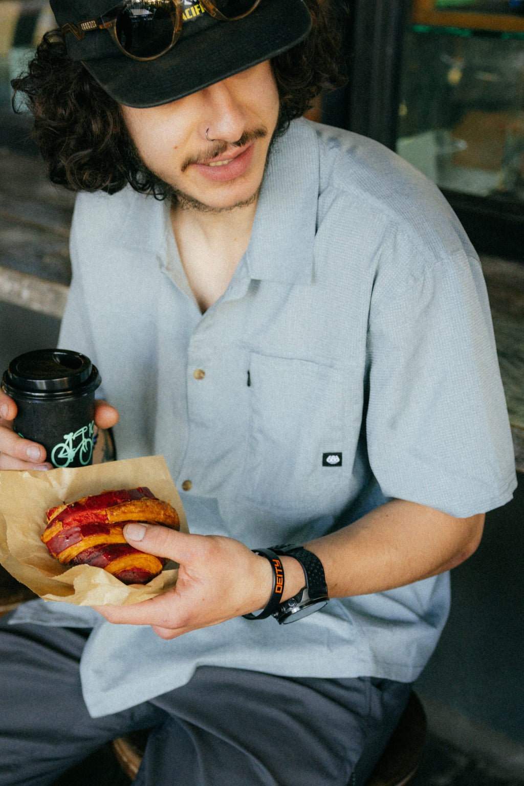 A person with curly hair and a mustache, wearing the 686 Mens Canopy Perforated Button Up by 686 and a black hat, sits outside holding a cup of coffee and a paper-wrapped sandwich.