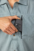 Wearing the 686 Mens Canopy Perforated Button Up, a person places a black smartphone with three camera lenses into the shirts zippered chest pocket.