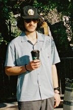 A man in sunglasses, a black cap, and the 686 Mens Canopy Perforated Button Up shirt stands outdoors with a coffee cup as leafy shadows from the sunlight fall across his 686 shirt.