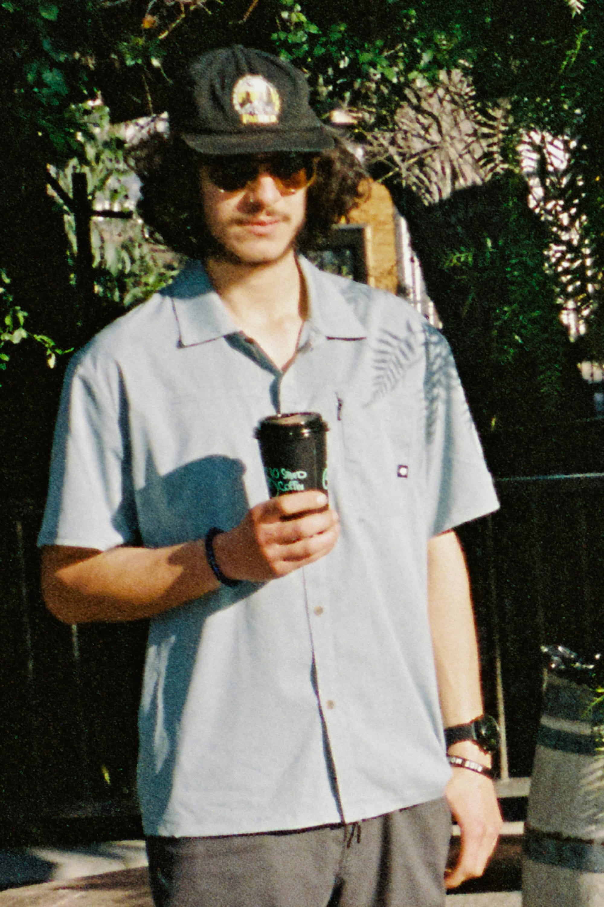 A man in sunglasses, a black cap, and the 686 Mens Canopy Perforated Button Up shirt stands outdoors with a coffee cup as leafy shadows from the sunlight fall across his 686 shirt.
