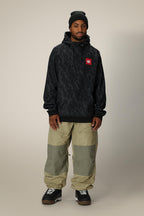 A man wearing a black beanie, 686 Mens Bonded Fleece Pullover Hoody by 686 with a red logo, light brown and olive cargo pants, and black sneakers stands against a plain beige background.