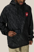 Wearing the 686 Mens Bonded Fleece Pullover Hoody, a person in khaki pants slips a black smartphone into its front pocket. The water-resistant tech hoodie features a red 686 logo and bold patterns, with their face partially out of frame.