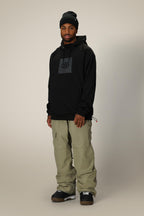 A man wearing the 686 Mens Bonded Fleece Pullover Hoody in black, featuring a chest graphic, paired with beige cargo pants and brown sneakers, stands against a plain light-colored background.
