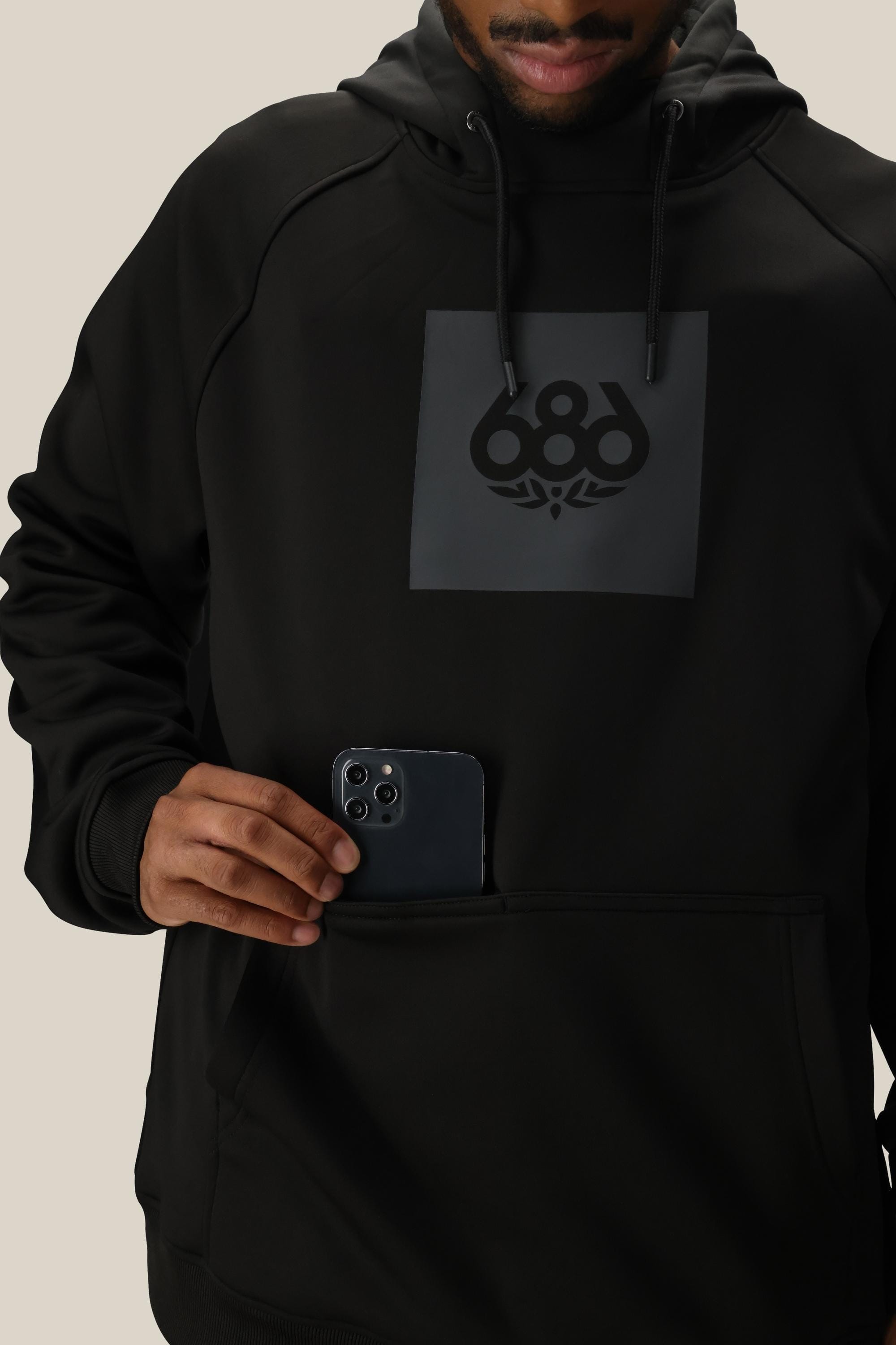 A person in a 686 Mens Bonded Fleece Pullover Hoody adds a black smartphone to the large front pocket. The hoodie is black with a front logo, and the background is plain and light-colored.