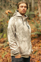 A man with light brown hair and a beard stands in a forest wearing the 686 Mens Bonded Fleece Pullover Hoody. This light gray and beige camo hoodie by 686 features fleece lining, ideal for cool autumn days outdoors.