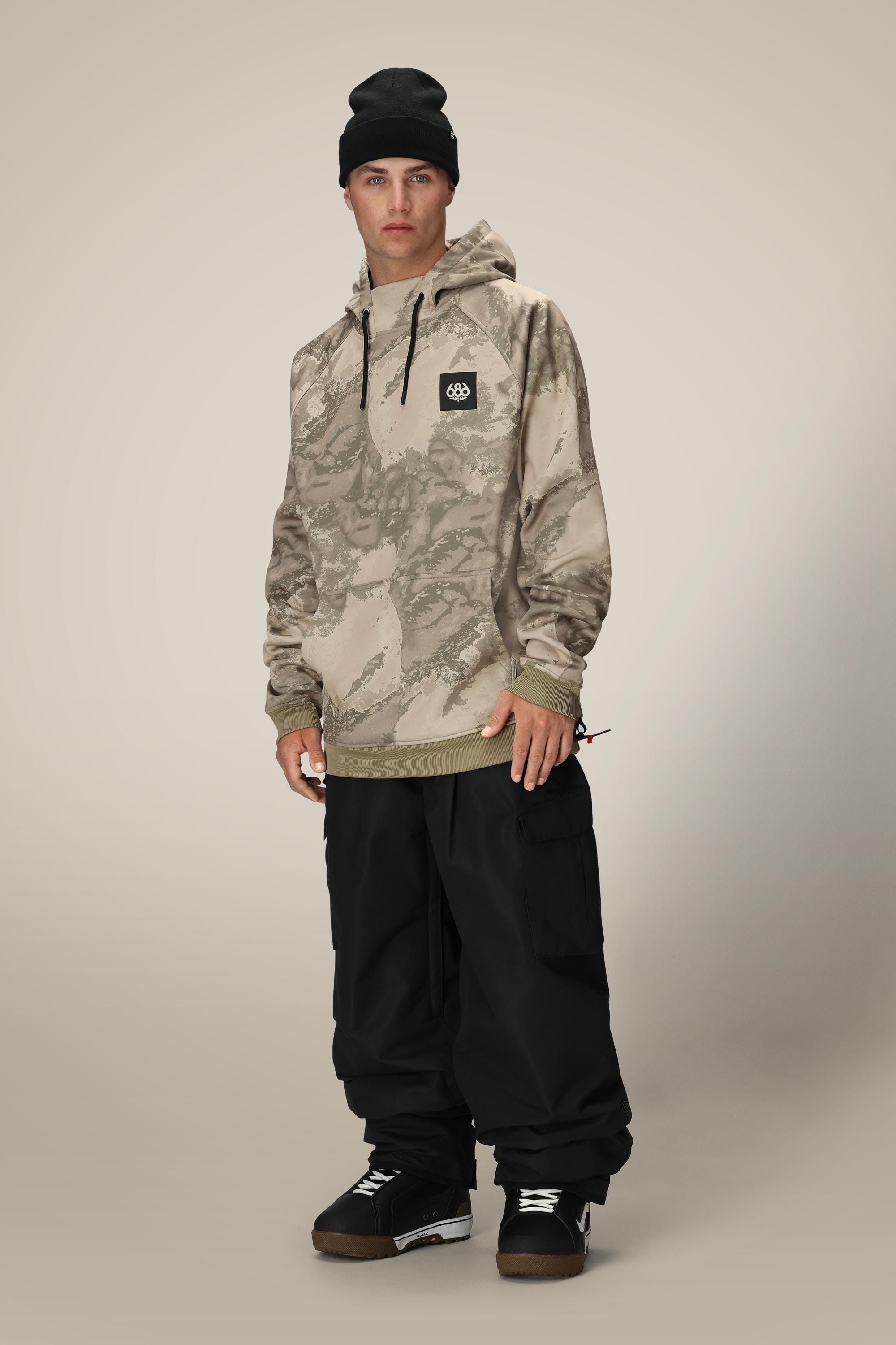 A person stands against a plain, light background wearing a black beanie, 686 Mens Bonded Fleece Pullover Hoody by 686 in beige camouflage with a logo patch, black cargo pants, and black boots.