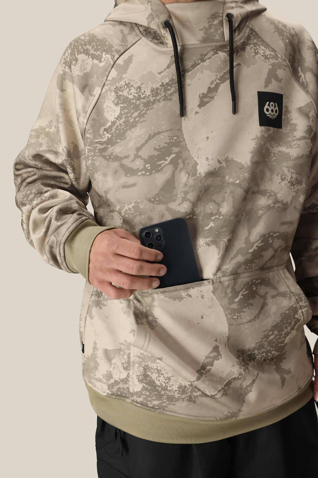A person, face not visible, wears the 686 Mens Bonded Fleece Pullover Hoody in beige camouflage with fleece lining and a black “686” logo, holding a black smartphone partly tucked into its water-resistant front pocket.