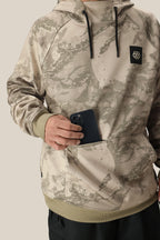 A person, face not visible, wears the 686 Mens Bonded Fleece Pullover Hoody in beige camouflage with fleece lining and a black “686” logo, holding a black smartphone partly tucked into its water-resistant front pocket.
