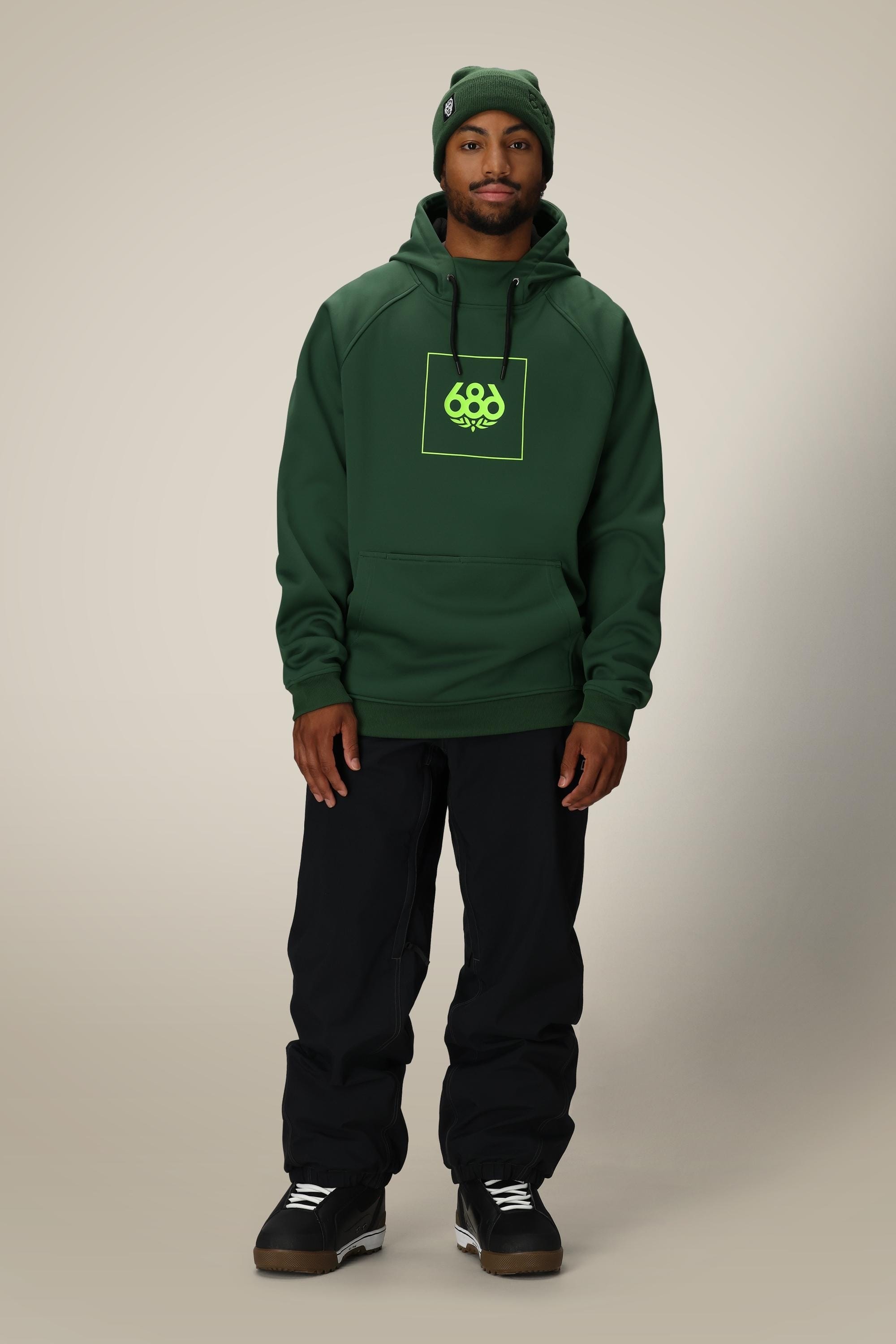 A man in a dark green beanie, black pants, black sneakers, and a 686 Mens Bonded Fleece Pullover Hoody with neon logo stands facing forward against a plain light background.
