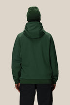 A person faces away from the camera wearing the 686 Mens Bonded Fleece Pullover Hoody in dark green, with a matching green beanie and black pants, standing against a plain beige background.