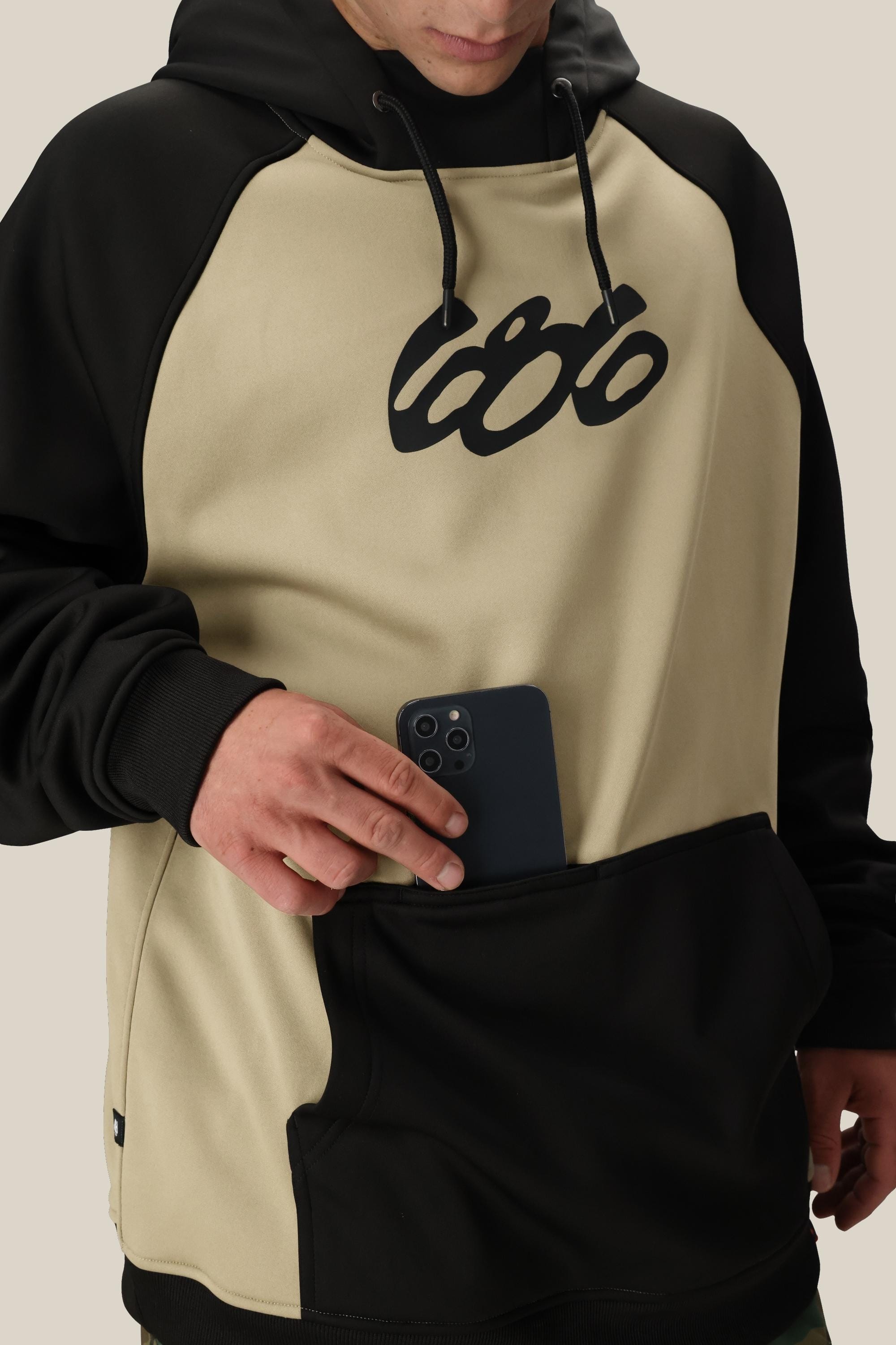 A person wearing the 686 Mens Bonded Fleece Pullover Hoody, featuring a two-tone beige and black design with cozy fleece lining, places a black smartphone into the front kangaroo pocket. The brand “686” is visible on the hoodie.