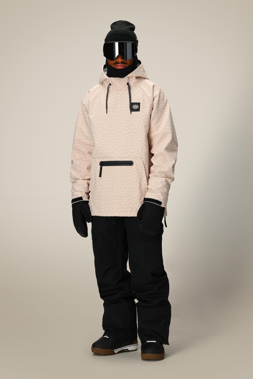 A person faces forward in a 686 Mens Waterproof Hyperchromic Pullover Hoody (beige, 10k waterproofing) with black snow pants, gloves, beanie, and ski goggles. The winter or snowboarding outfit stands against a plain gray background.