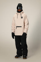 A person faces forward in a 686 Mens Waterproof Hyperchromic Pullover Hoody (beige, 10k waterproofing) with black snow pants, gloves, beanie, and ski goggles. The winter or snowboarding outfit stands against a plain gray background.