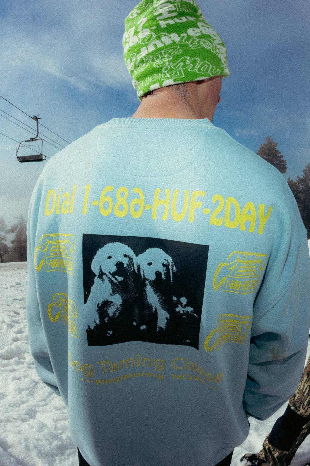 A person in a green beanie and blue 686 HUF Premium Heavyweight Crew Sweatshirt stands in the snow by a ski lift. The cotton polyester spandex blend sweatshirt features yellow dog graphics and “Dial 1-888-HUF-2DAY” on the back.