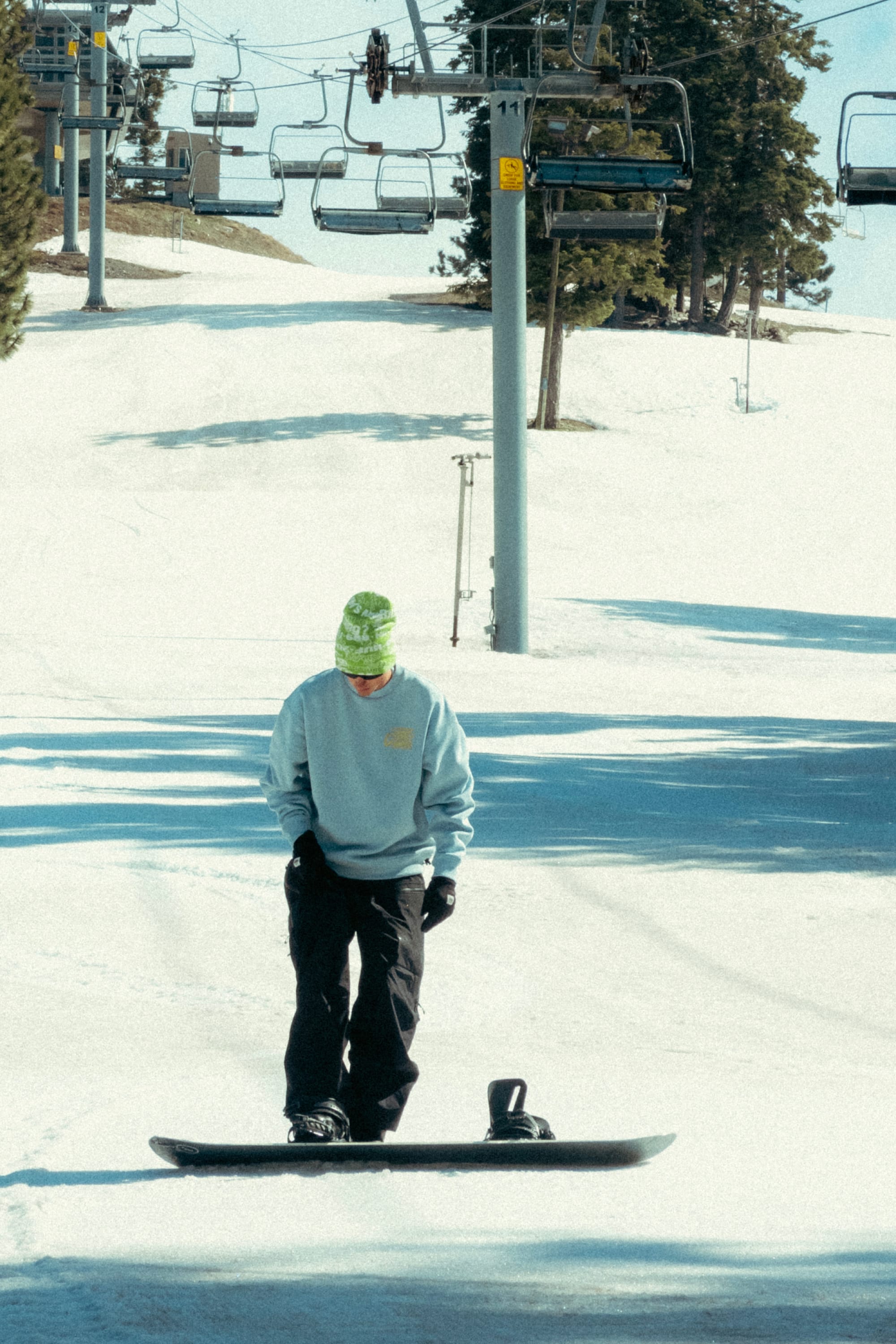 Wearing a green beanie and the 686 HUF Premium Heavyweight Crew Sweatshirt, a person stands on a snowboard atop a snowy slope with empty ski lifts and trees in the background.