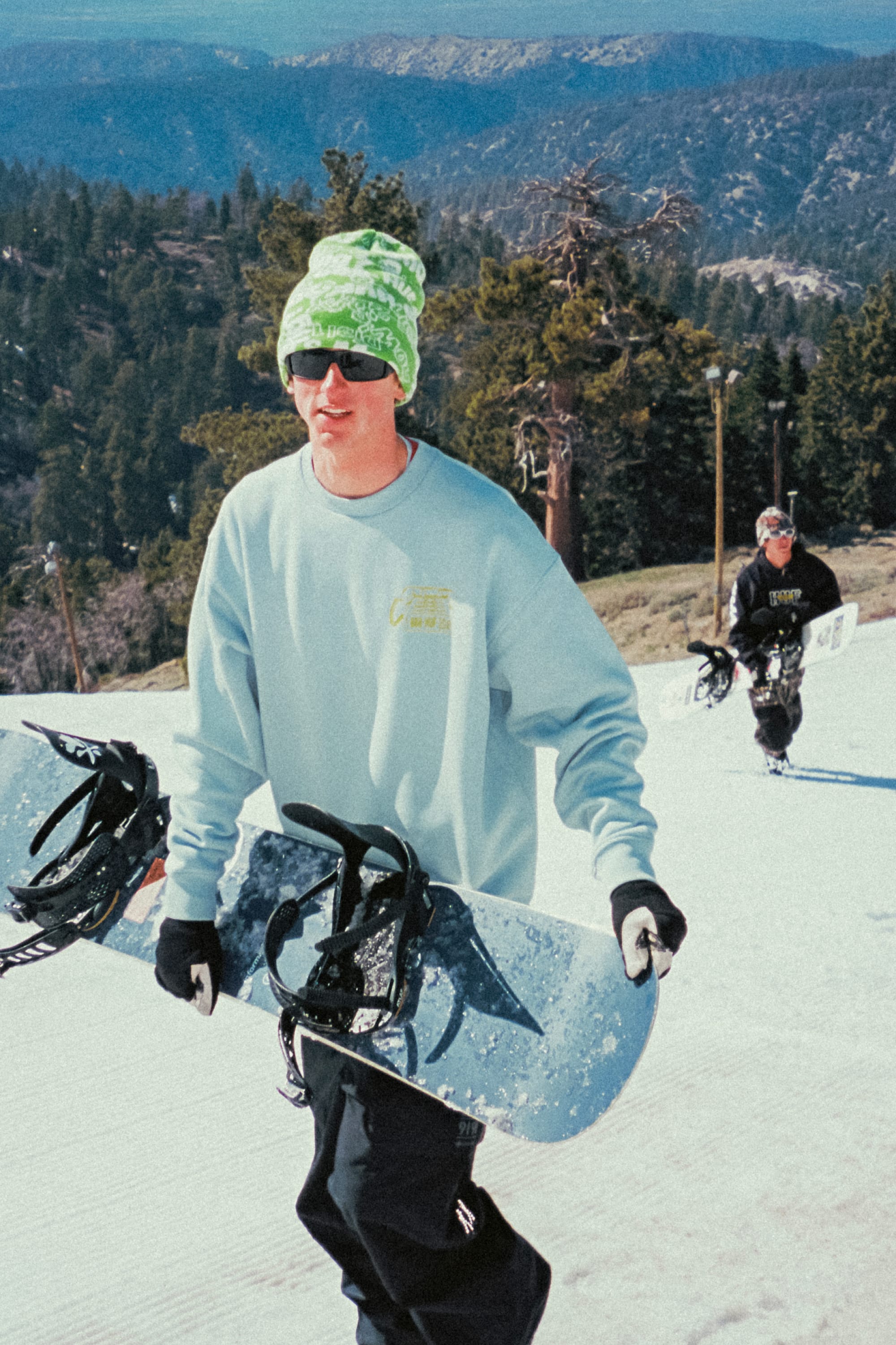 Wearing a green beanie, sunglasses, and the 686 HUF Premium Heavyweight Crew Sweatshirt, a man carries a snowboard up a snowy slope with another snowboarder following, pine trees and mountains in the background.