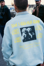 A man outdoors among a small group wears a light blue 686 HUF Premium Heavyweight Crew Sweatshirt with yellow Dog Taming Classes text, graphics of two dogs, and a phone number. Brand: 686.