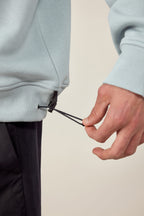 A person tightens the drawstring at the hem of a light blue 686 HUF Premium Heavyweight Crew Sweatshirt, highlighting the lower part of this cotton polyester spandex blend by 686 and their hand in focus.