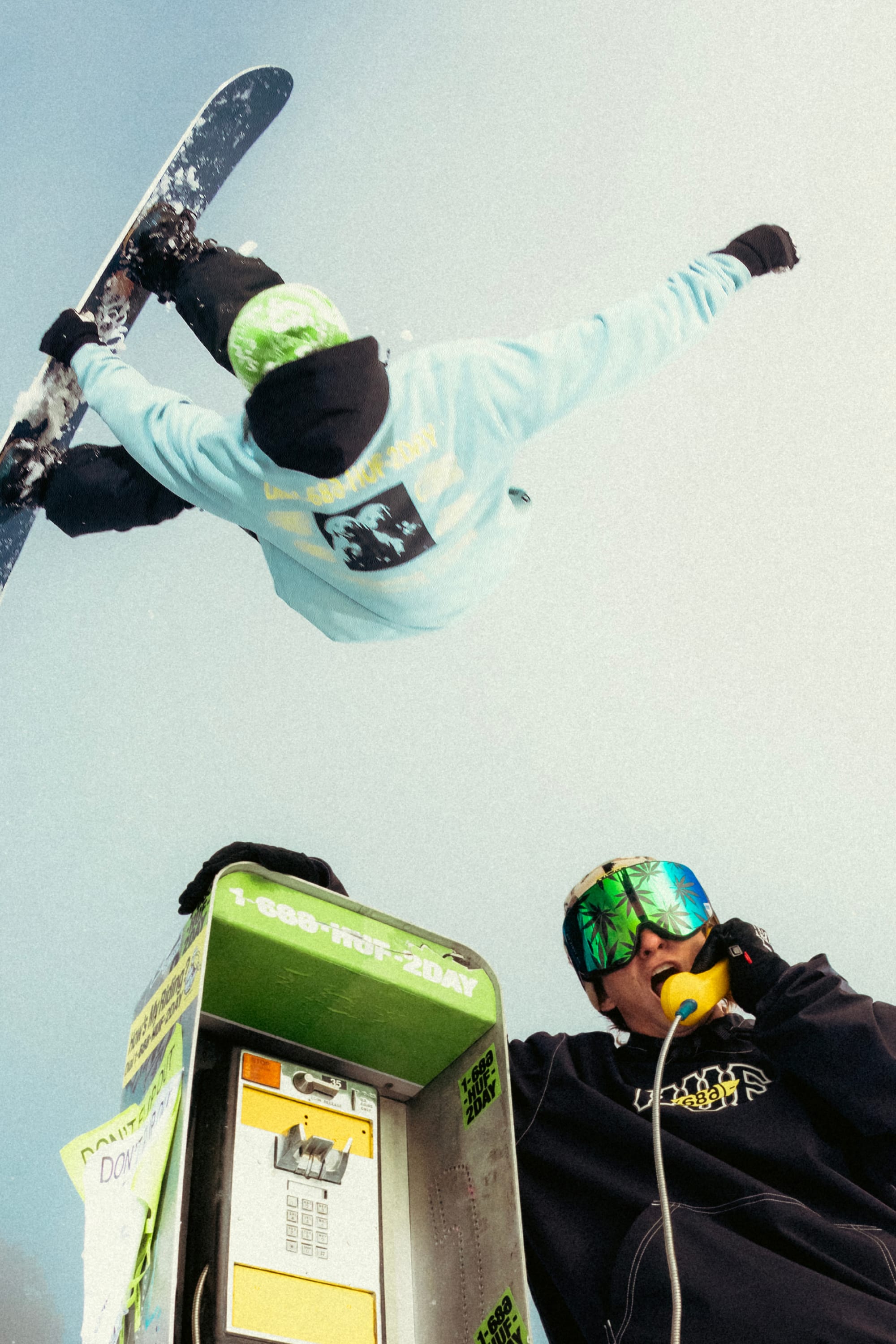 Wearing the 686 HUF Premium Heavyweight Crew Sweatshirt, a snowboarder soars above a payphone as another person in ski goggles uses the phone, both set against a vivid snowy backdrop.