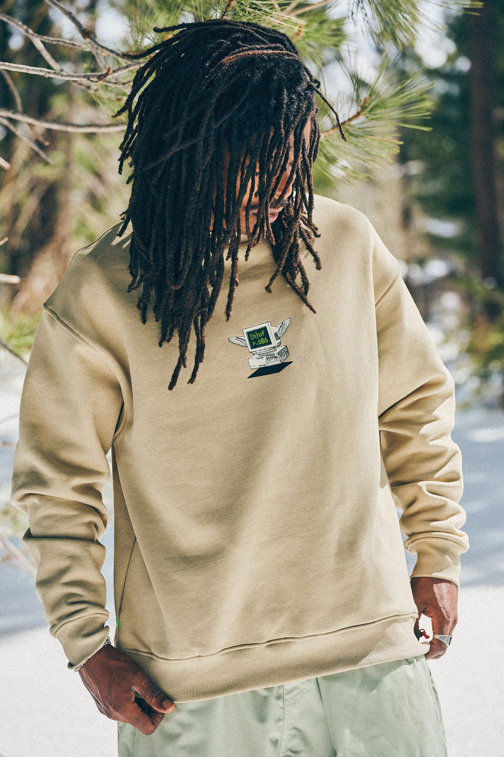 A person with long dreadlocks stands outdoors near pine trees and snow, wearing the 686 HUF Premium Heavyweight Crew Sweatshirt by 686 with hands in their pockets, looking down.