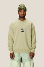 A man wearing a beige 686 HUF Premium Heavyweight Crew Sweatshirt by 686, mint green pants, and a green patterned beanie stands facing forward against a plain light background.