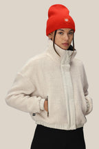 A person wears the 686 Womens Outpost Sherpa Jacket in white with zippered pockets and a red beanie, standing against a plain beige background and looking slightly to the side.