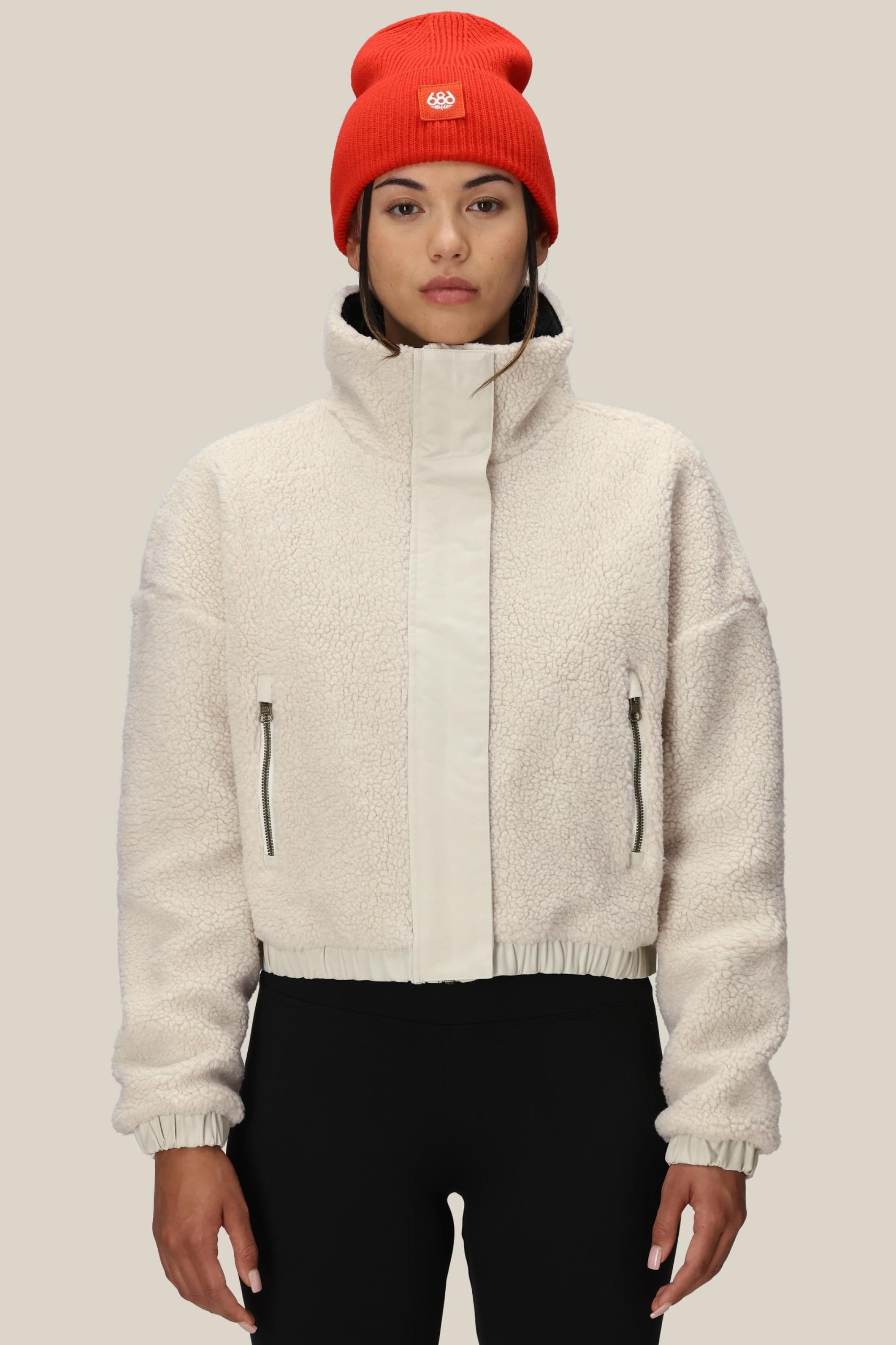 A woman wears the 686 Womens Outpost Sherpa Jacket in cream, featuring a high collar and zippered pockets, with black pants and a bright red beanie, standing against a plain light beige background.