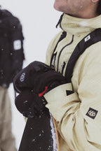 A person in a light jacket wears 686 Men's GORE-TEX Leather Linear™ Mitts and holds a snowboard. Their face is partially out of frame with visible snowflakes, while someone in dark clothing is blurred in the background.