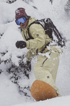 A snowboarder in beige outerwear, a maroon beanie, blue goggles, and 686 Men's GORE-TEX Leather Linear™ Mitts rides through fresh snow on a mountainside amid snow-covered trees.