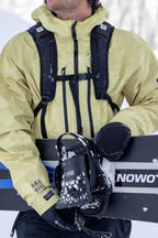 A person holds a snowboard vertically, wearing a yellow jacket and 686 Men's GORE-TEX Leather Linear™ Mitts by 686. Only the bottom of their face is visible amid snow and winter gear, highlighting a cold outdoor setting.