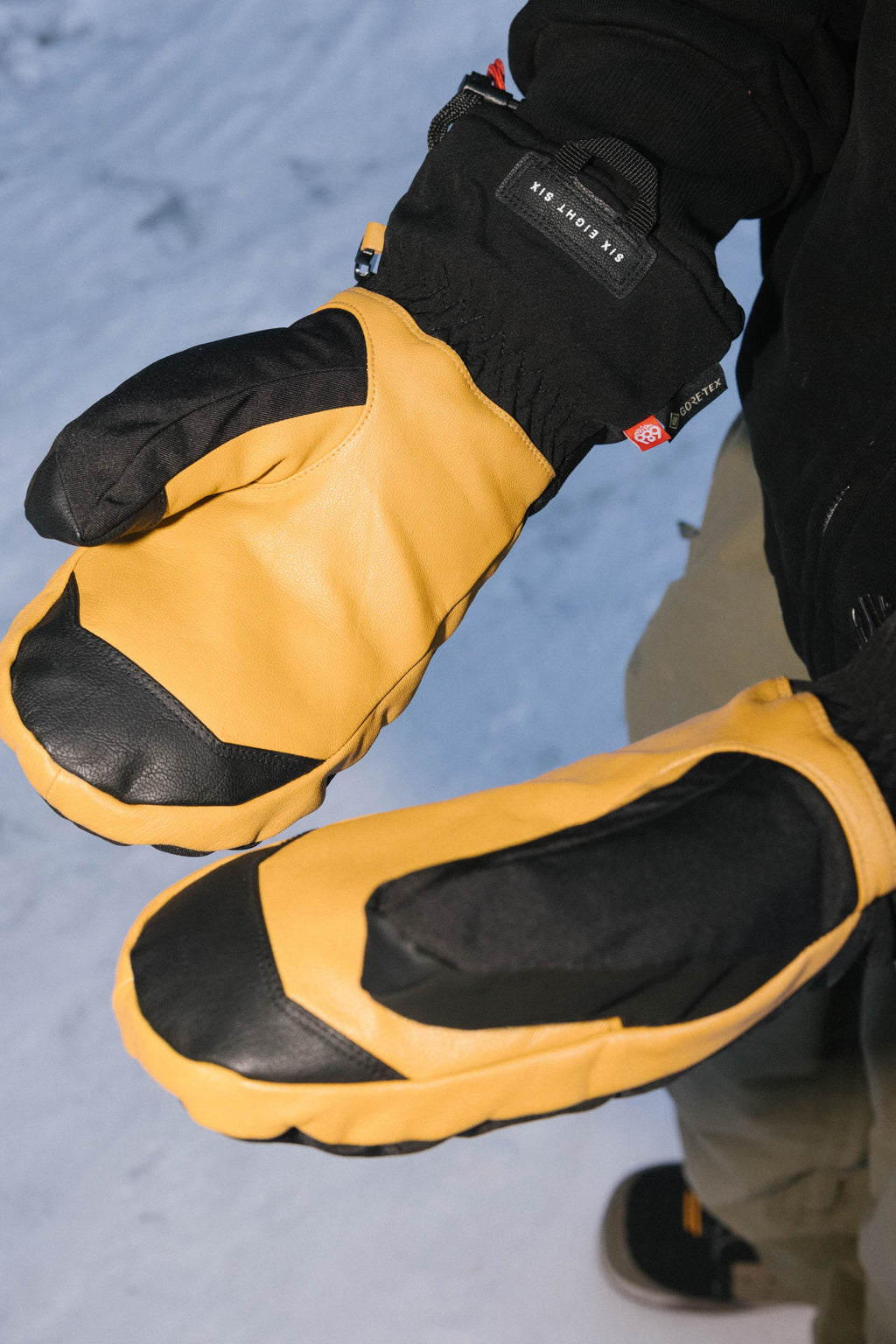 A person wearing 686 Men's GORE-TEX Leather Linear™ Mitts with leather palms and yellow backs stands on snow, arms extended. These 686 mitts provide durable grip with black accents and GORE-TEX protection for winter activities.