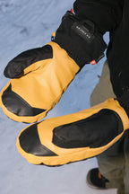 A person wearing 686 Men's GORE-TEX Leather Linear™ Mitts with leather palms and yellow backs stands on snow, arms extended. These 686 mitts provide durable grip with black accents and GORE-TEX protection for winter activities.