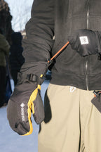Wearing a black jacket, a person stands in the snow using an orange zipper pull on their sleeve while wearing 686 Men's GORE-TEX Leather Linear™ Mitts in black and yellow by 686, featuring a durable leather palm.