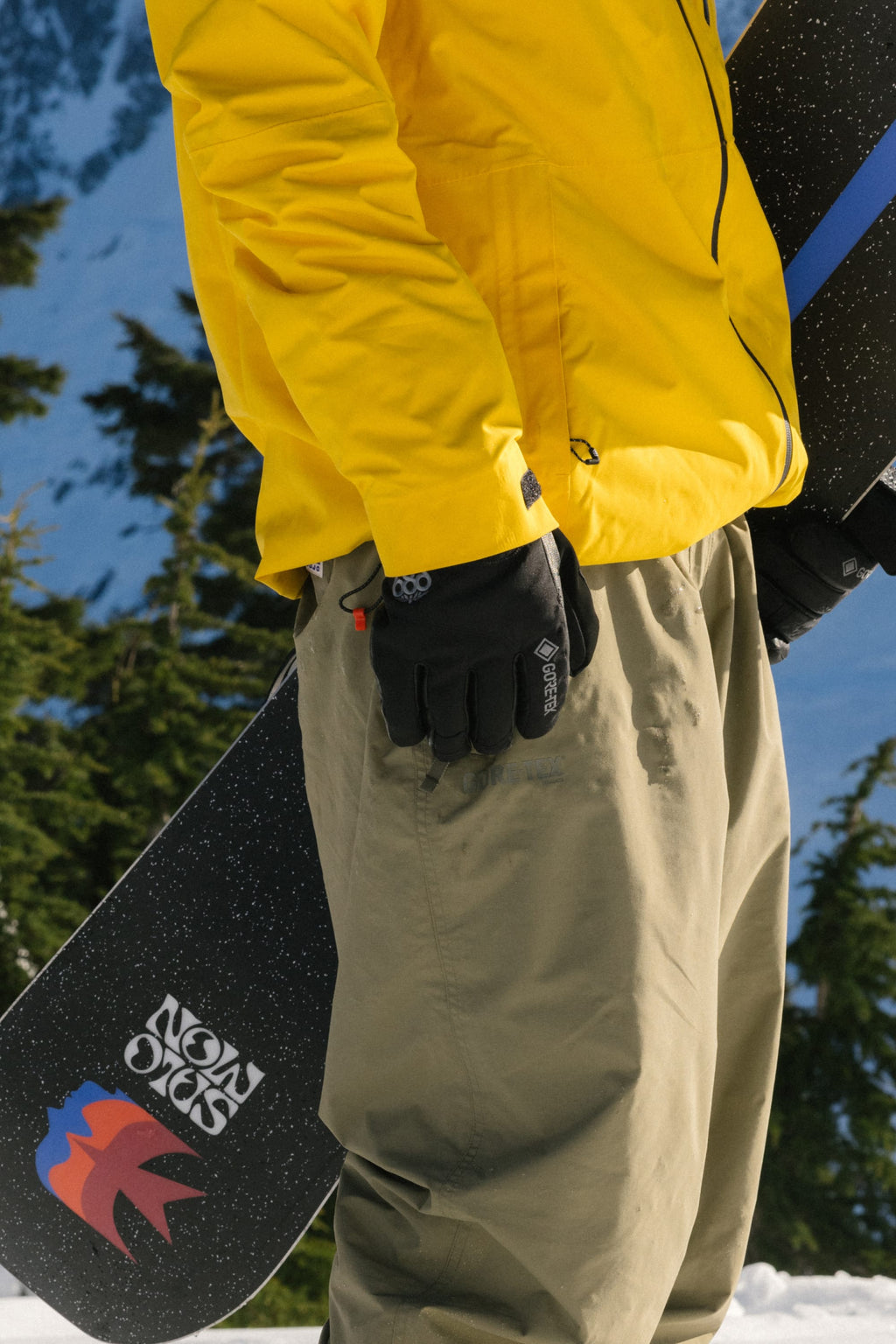 Wearing a yellow jacket and olive-green pants, a person stands in the snow holding a snowboard with a mountain logo, sporting 686 Men's GORE-TEX Leather Linear™ Gloves for extra grip, surrounded by evergreen trees and snowy mountains.