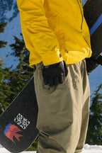Wearing a yellow jacket and olive-green pants, a person stands in the snow holding a snowboard with a mountain logo, sporting 686 Men's GORE-TEX Leather Linear™ Gloves for extra grip, surrounded by evergreen trees and snowy mountains.