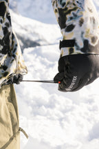 Wearing patterned winter clothing, a person adjusts a jacket drawstring in the snow while wearing black 686 Men's GORE-TEX Leather Linear™ Gloves.