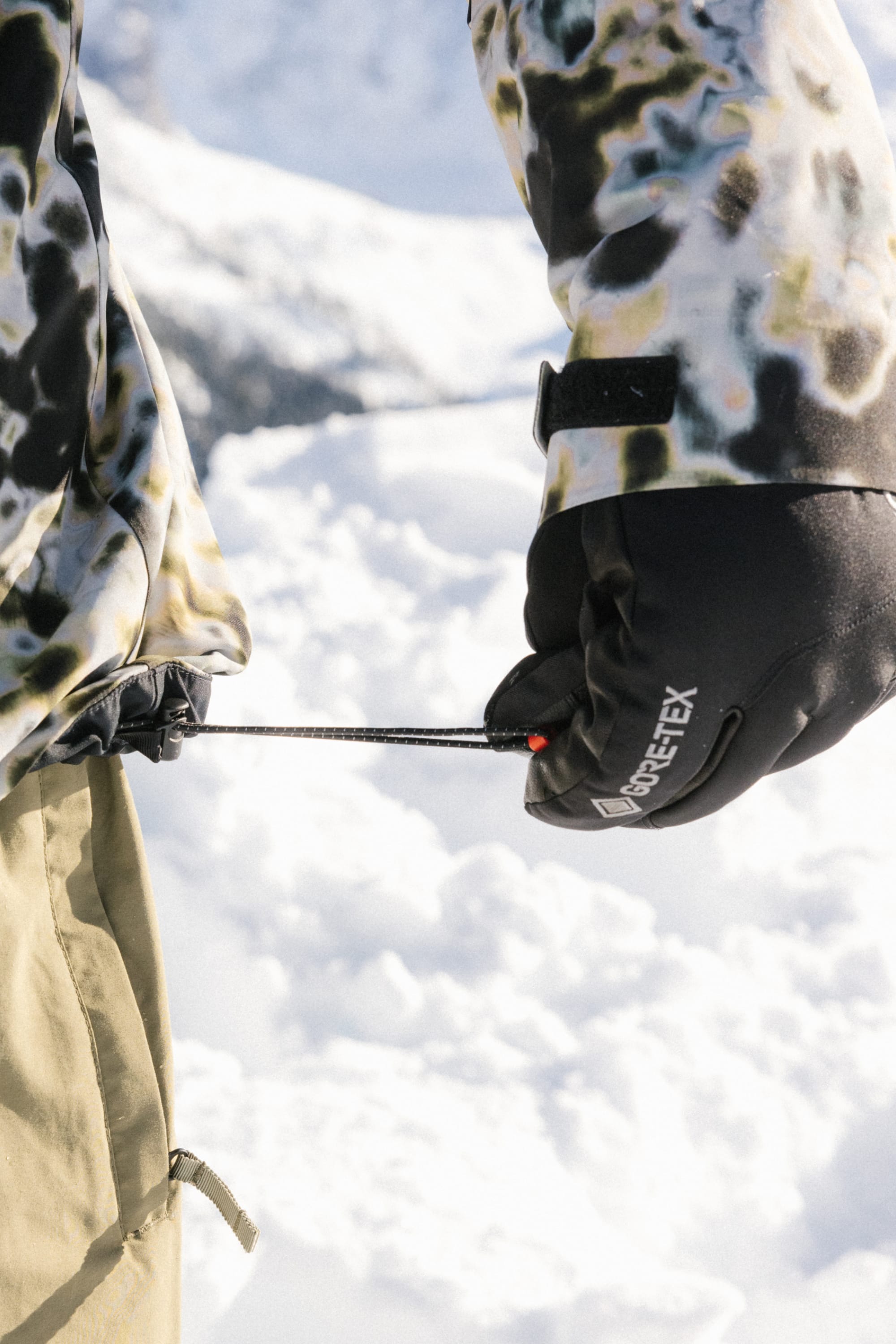 Wearing patterned winter clothing, a person adjusts a jacket drawstring in the snow while wearing black 686 Men's GORE-TEX Leather Linear™ Gloves.