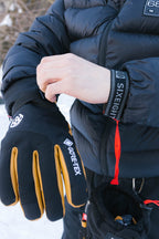 A person in a black puffer jacket puts on a 686 Men's GORE-TEX Leather Linear™ Glove, featuring a leather palm. The other glove, attached by a red strap, hangs from their wrist with snow in the background.