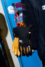 A pair of 686 Men's GORE-TEX Leather Linear™ Gloves in black and tan with logo details and a durable leather palm hang from a blue locker door with stickers. A black jacket is partially visible beside the gloves.