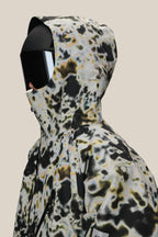 A person wearing the 686 Men's Hydra™ Thermagraph® Jacket by 686, featuring a black, white, and yellow abstract pattern, paired with large black visor sunglasses and a black face covering against a plain beige background.