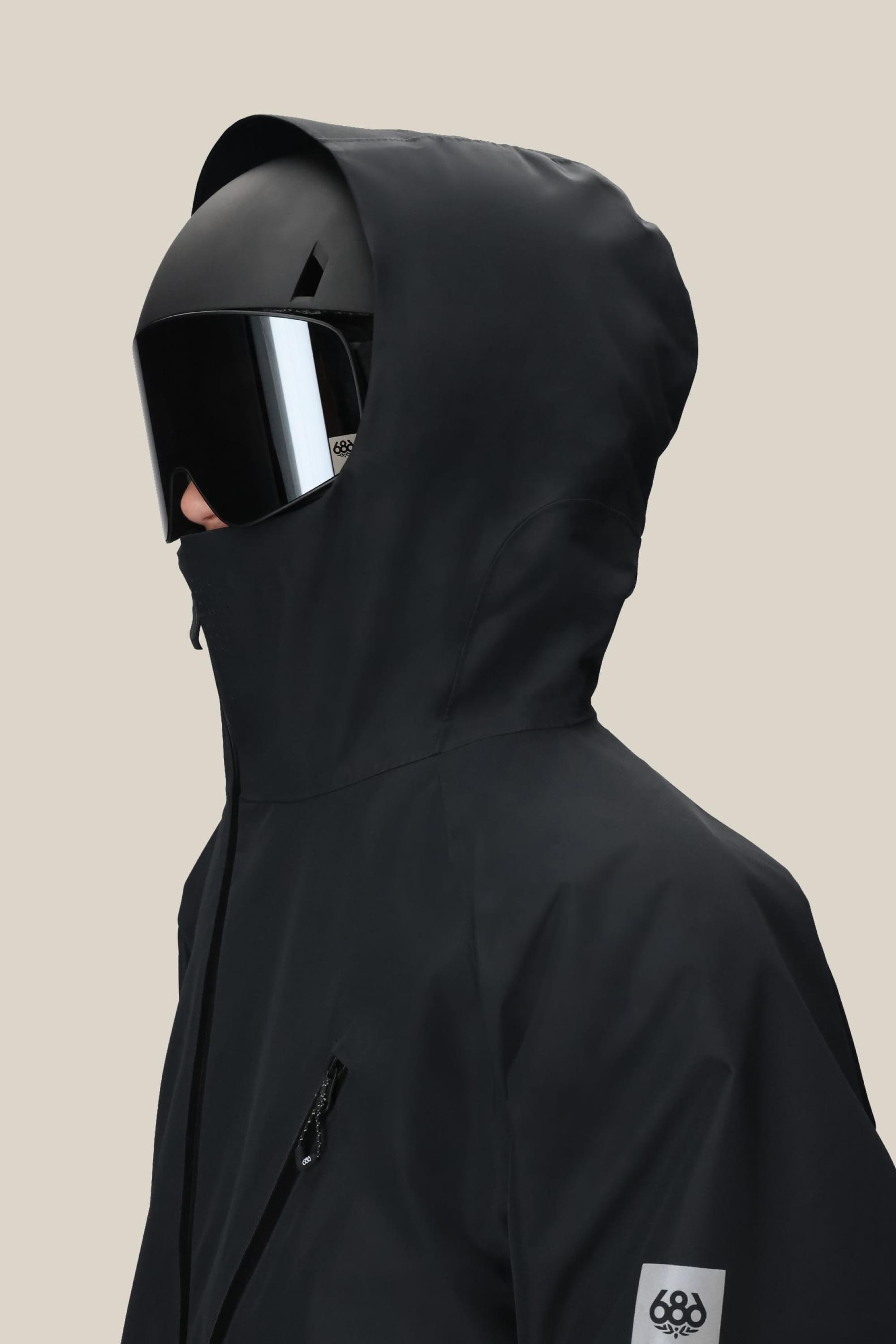A man in the 686 Men's Hydra™ Thermagraph® Jacket with a high collar, black ski helmet, and reflective goggles stands in profile against a plain beige background.