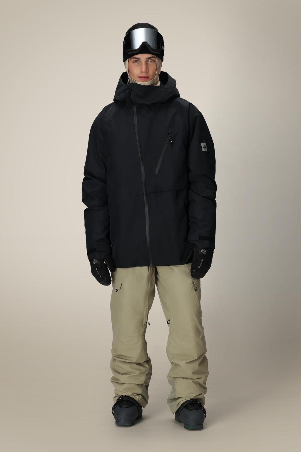 A person wears the 686 Men's Hydra™ Thermagraph® Jacket in black, paired with beige snow pants, black gloves, and a black helmet with reflective ski goggles, standing against a plain light background.