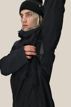 Wearing the 686 Men's Hydra™ Thermagraph® Jacket, a person in black unzips the underarm vent to show the body-mapped lining. They pair it with a beanie and layer a beige balaclava underneath for extra warmth.