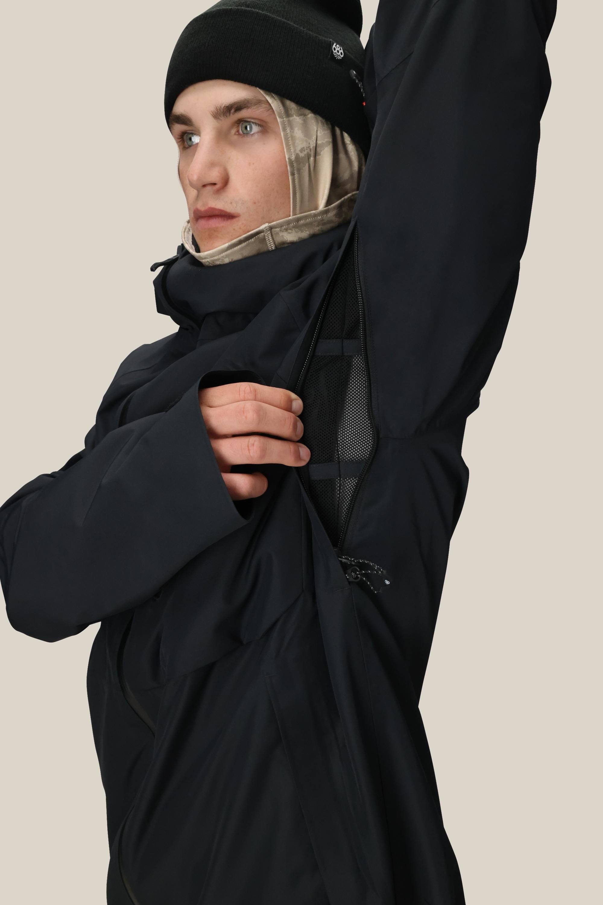 Wearing the 686 Men's Hydra™ Thermagraph® Jacket, a person in black unzips the underarm vent to show the body-mapped lining. They pair it with a beanie and layer a beige balaclava underneath for extra warmth.