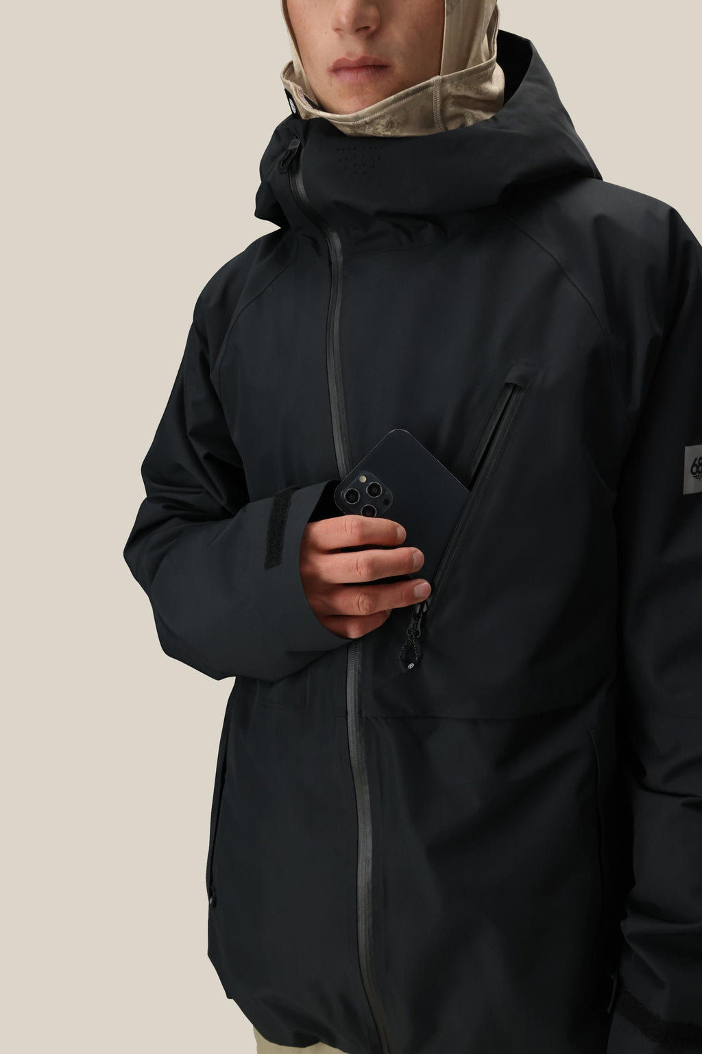 A person wearing the 686 Men's Hydra™ Thermagraph® Jacket, featuring a black hood, slips a smartphone into its zippered chest pocket; only their lower face shows against a plain light-colored background.