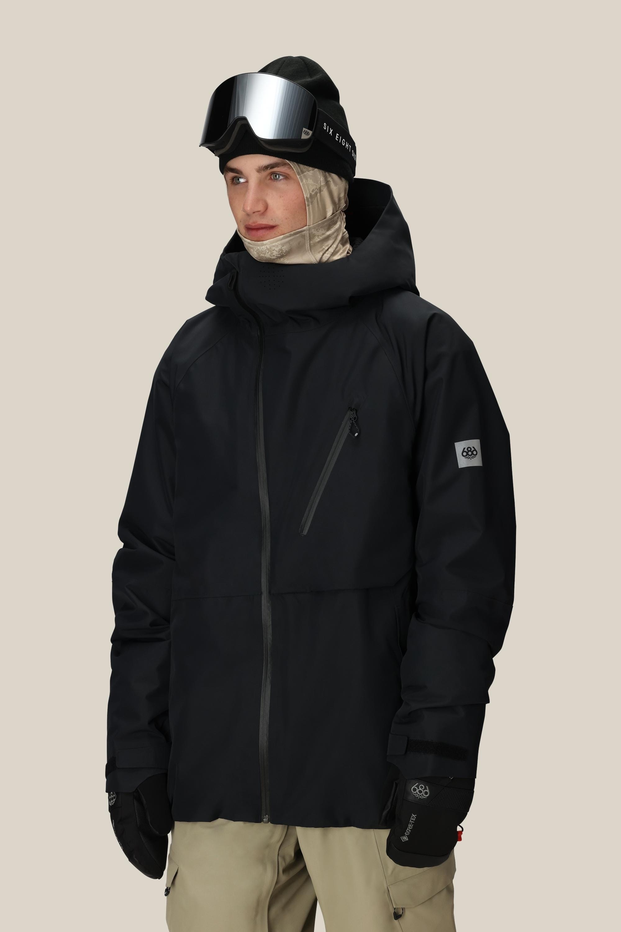 A person wears the 686 Men's Hydra™ Thermagraph® Jacket with a beige balaclava, black ski gloves, black beanie, and mirrored ski goggles, standing against a plain light background.