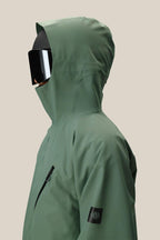 A person wearing the 686 Men's Hydra™ Thermagraph® Jacket, a green bestseller with a black sleeve patch and zippered chest pocket, faces left against a beige background. This 686 jacket blends practical style and comfort for any occasion.