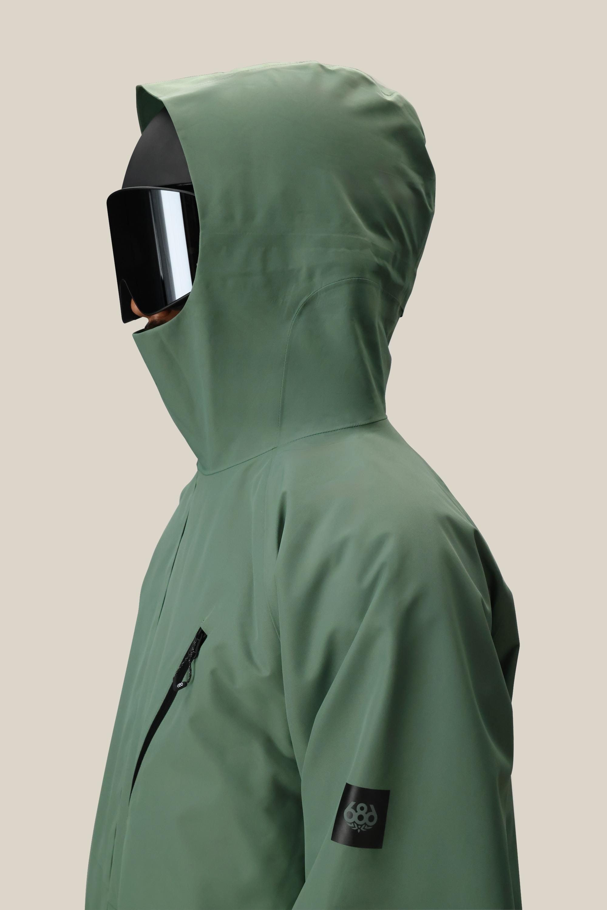 A person wearing the 686 Men's Hydra™ Thermagraph® Jacket, a green bestseller with a black sleeve patch and zippered chest pocket, faces left against a beige background. This 686 jacket blends practical style and comfort for any occasion.