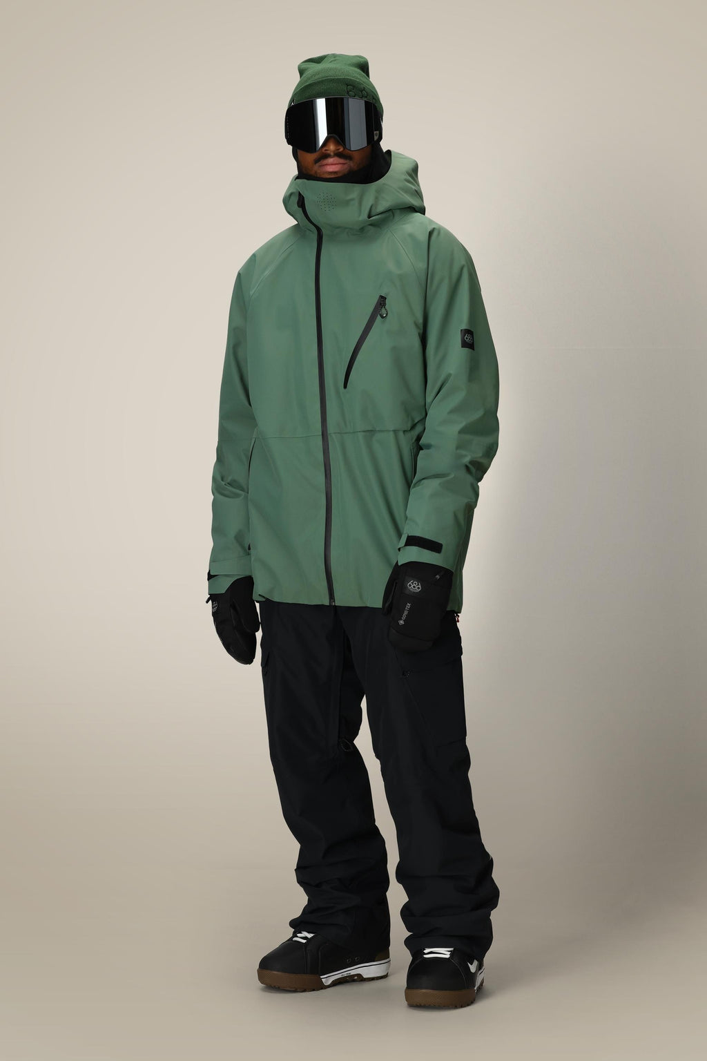 A person wears a 686 Men's Hydra™ Thermagraph® Jacket, black snow pants, black gloves, brown snow boots, a green beanie, and large reflective ski goggles against a plain light background.