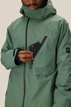 A person in a green 686 Men's Hydra™ Thermagraph® Jacket with body-mapped lining places a black smartphone into the partially unzipped chest pocket, their face partly visible against a plain beige background.